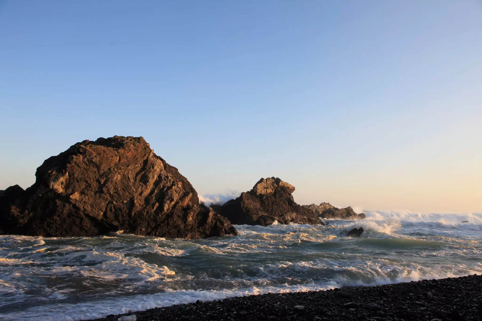 Waves crashing on large ocean rocks close to a rocky shore on a mostly sunny day.