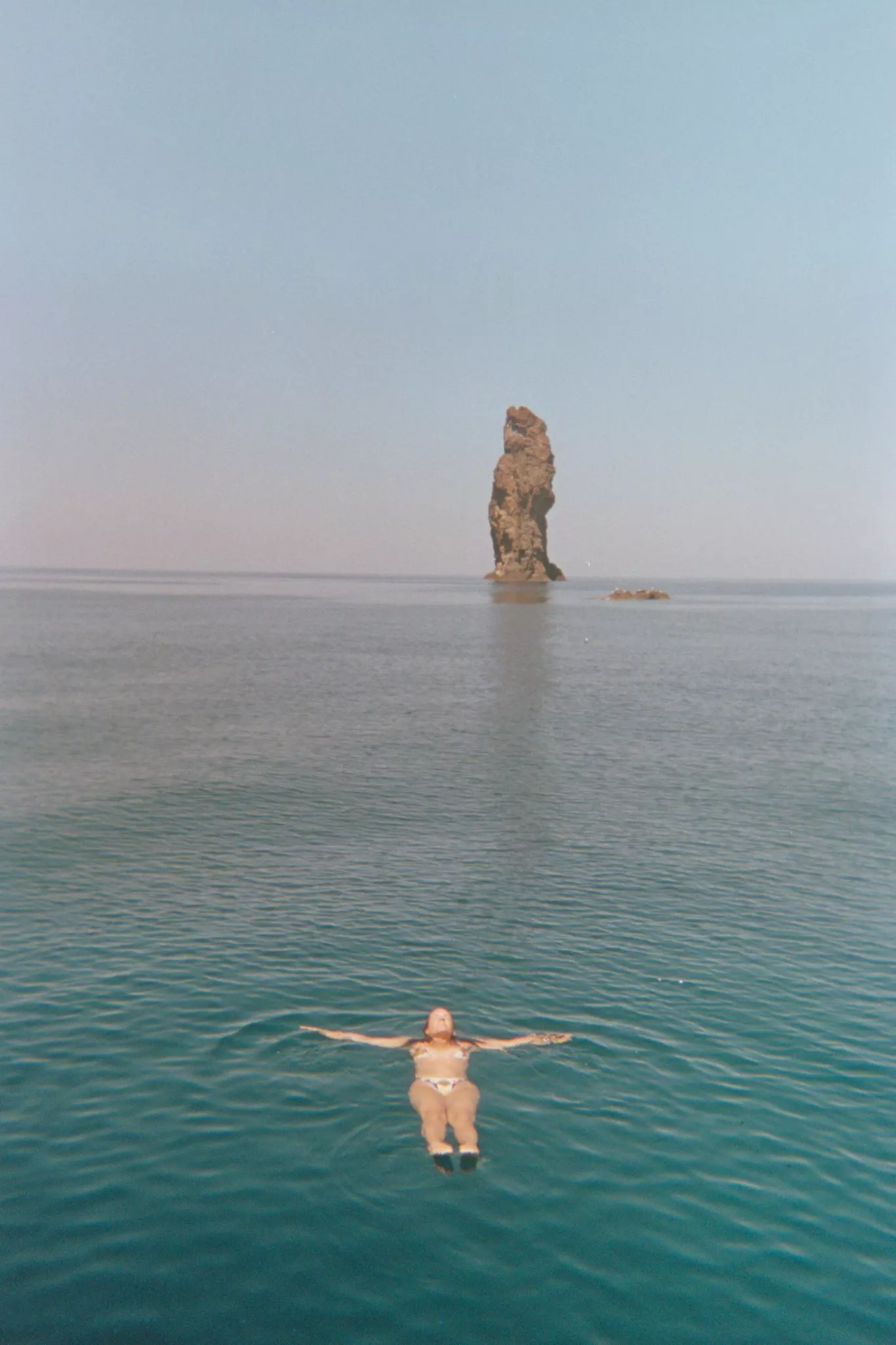 A woman floating on her back in the water with blue sky and a rock promontory behind her