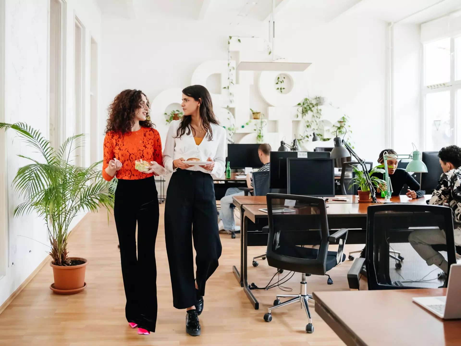 A transgender woman walking past some computer desks heading for her lunch with an office colleague