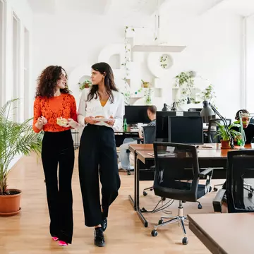 A transgender woman walking past some computer desks heading for her lunch with an office colleague