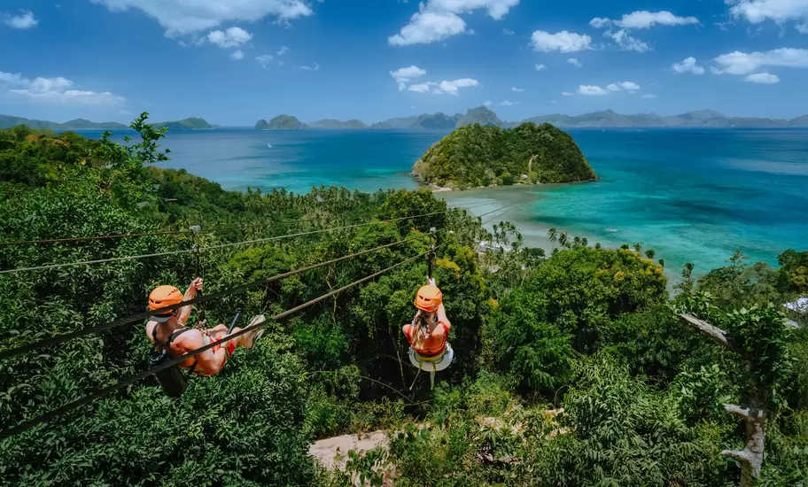 Two people in orange helmets ziplining over jungle toward a turquoise bay and islets near El Nido, Philippines