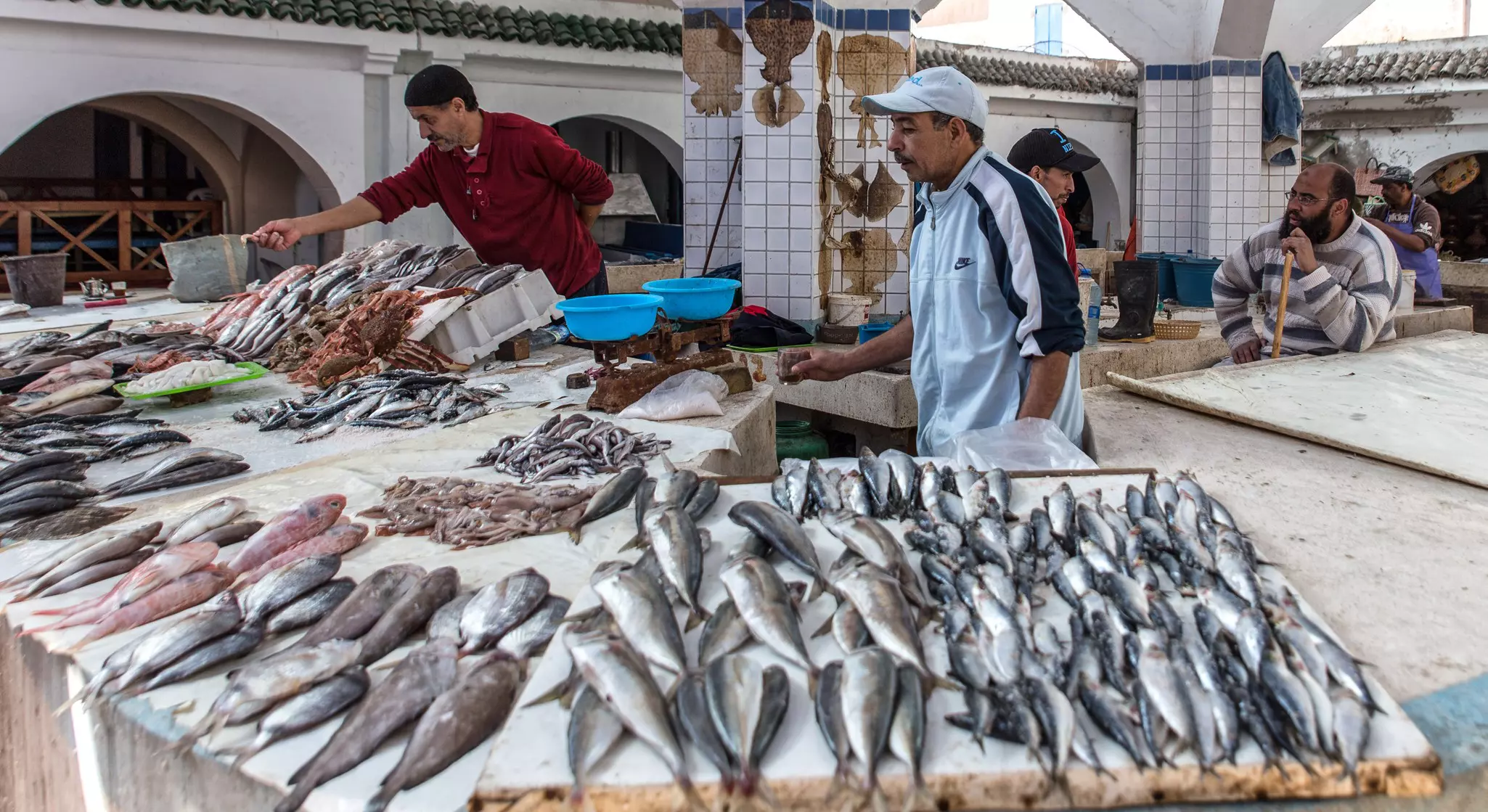 Two men stand behind a stall with fresh fish for sale in a food market.