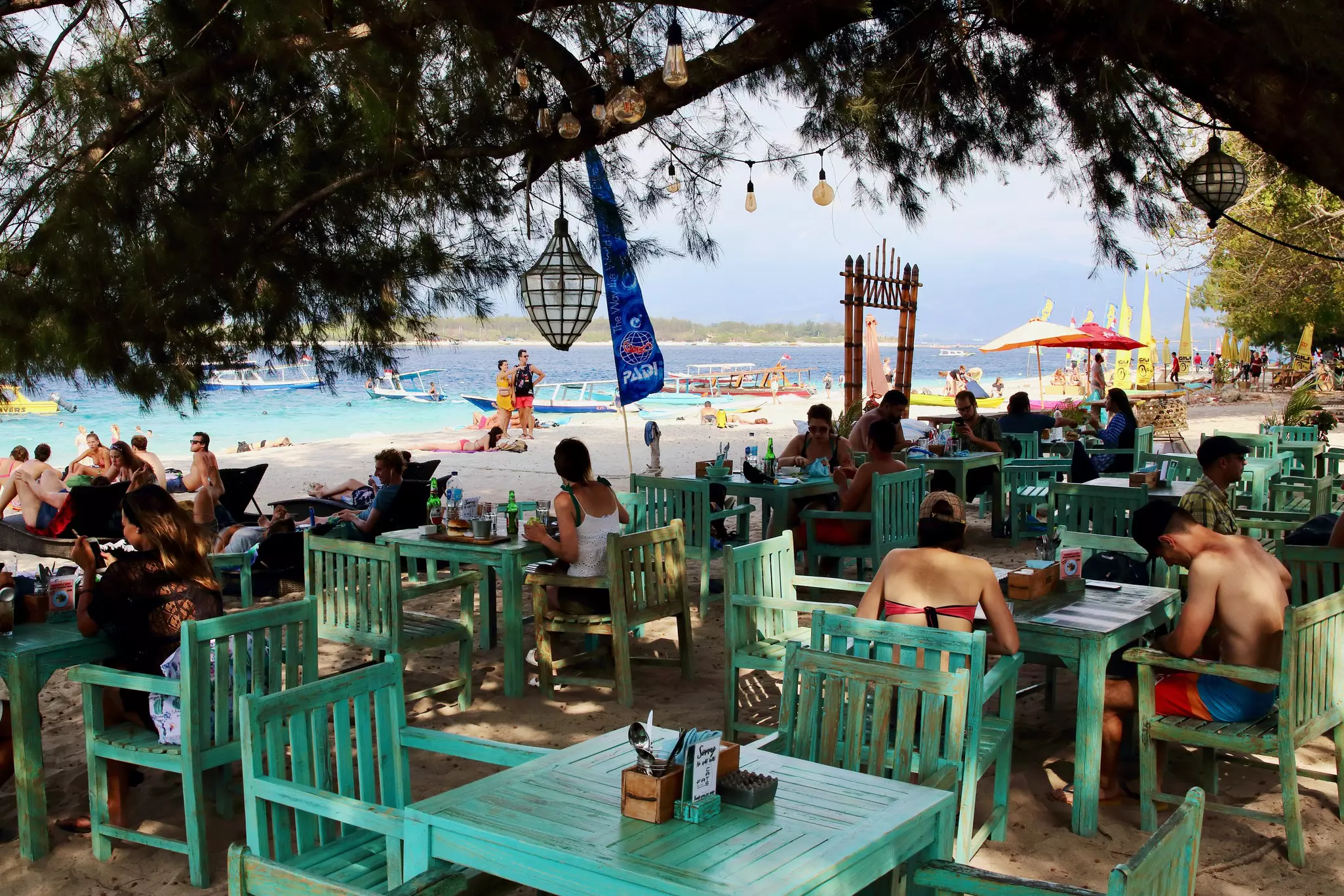 People in bathing suits sit at green-colored chairs and tables at a bar on the beach. Boats are visible in the water offshore.