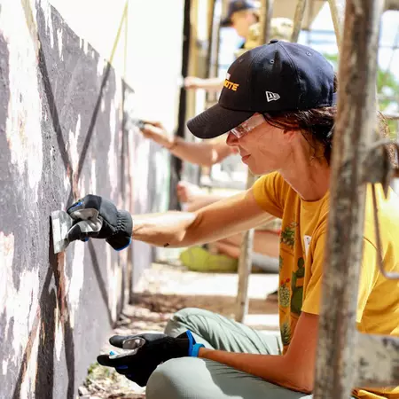 A woman in a yellow T-shirt and black baseball cap helps paint an exterior wall on a sunny day.