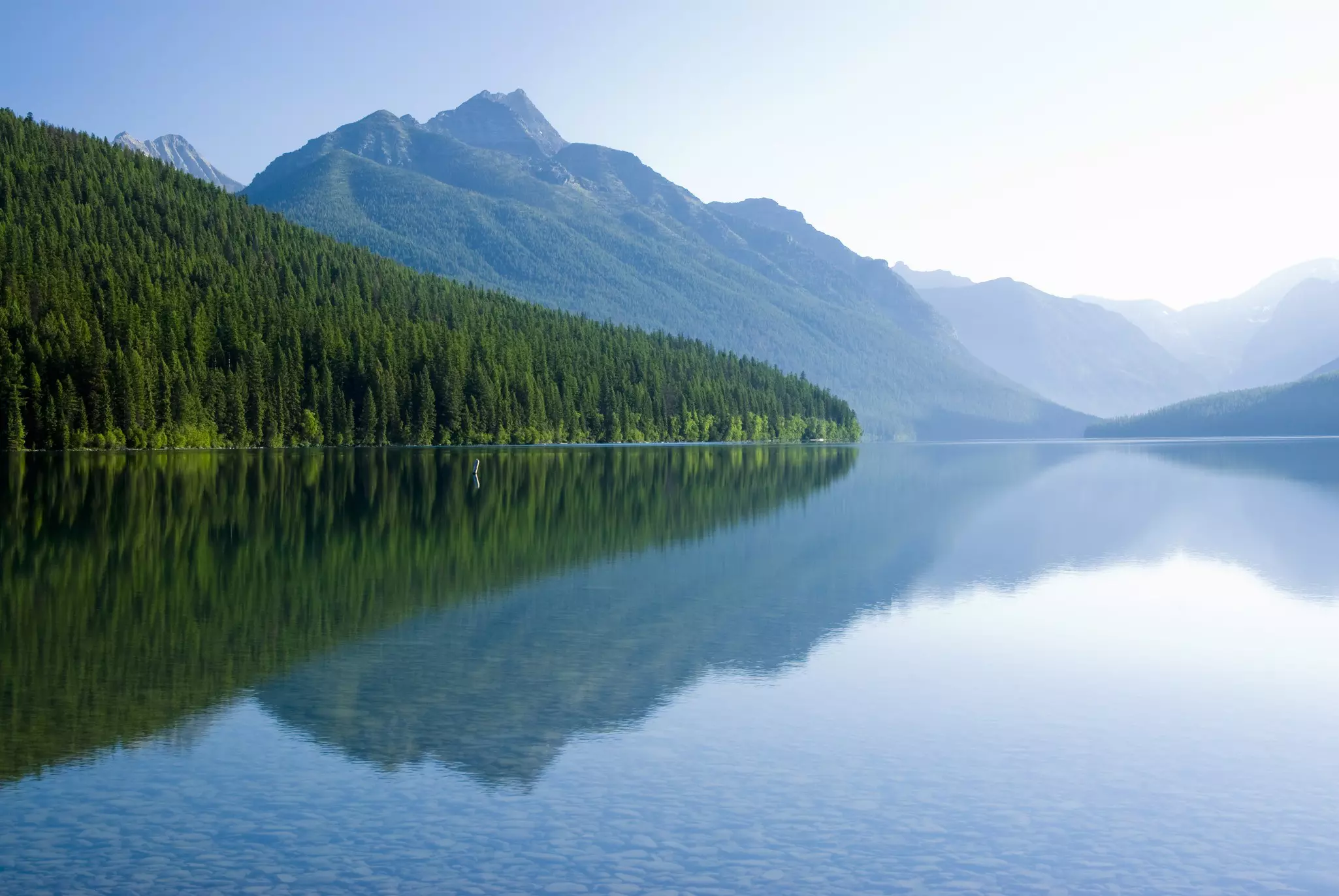 Mountains and trees reflected in a very still blue lake.