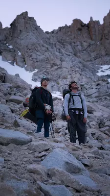 Two male hikers carrying backpacks pause on a mountain trail to watch the sunrise.