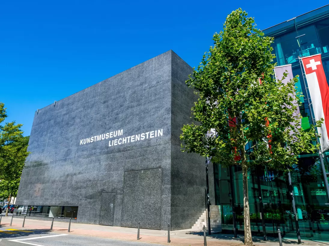 A windowless black polished-concrete cube lettered KUNSTMUSEUM LIECHTENSTEIN, beside a glass wing with flags flying, under a clear blue sky.