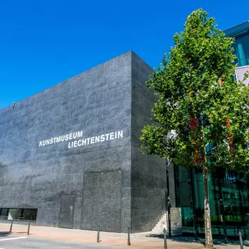 A windowless black polished-concrete cube lettered KUNSTMUSEUM LIECHTENSTEIN, beside a glass wing with flags flying, under a clear blue sky.