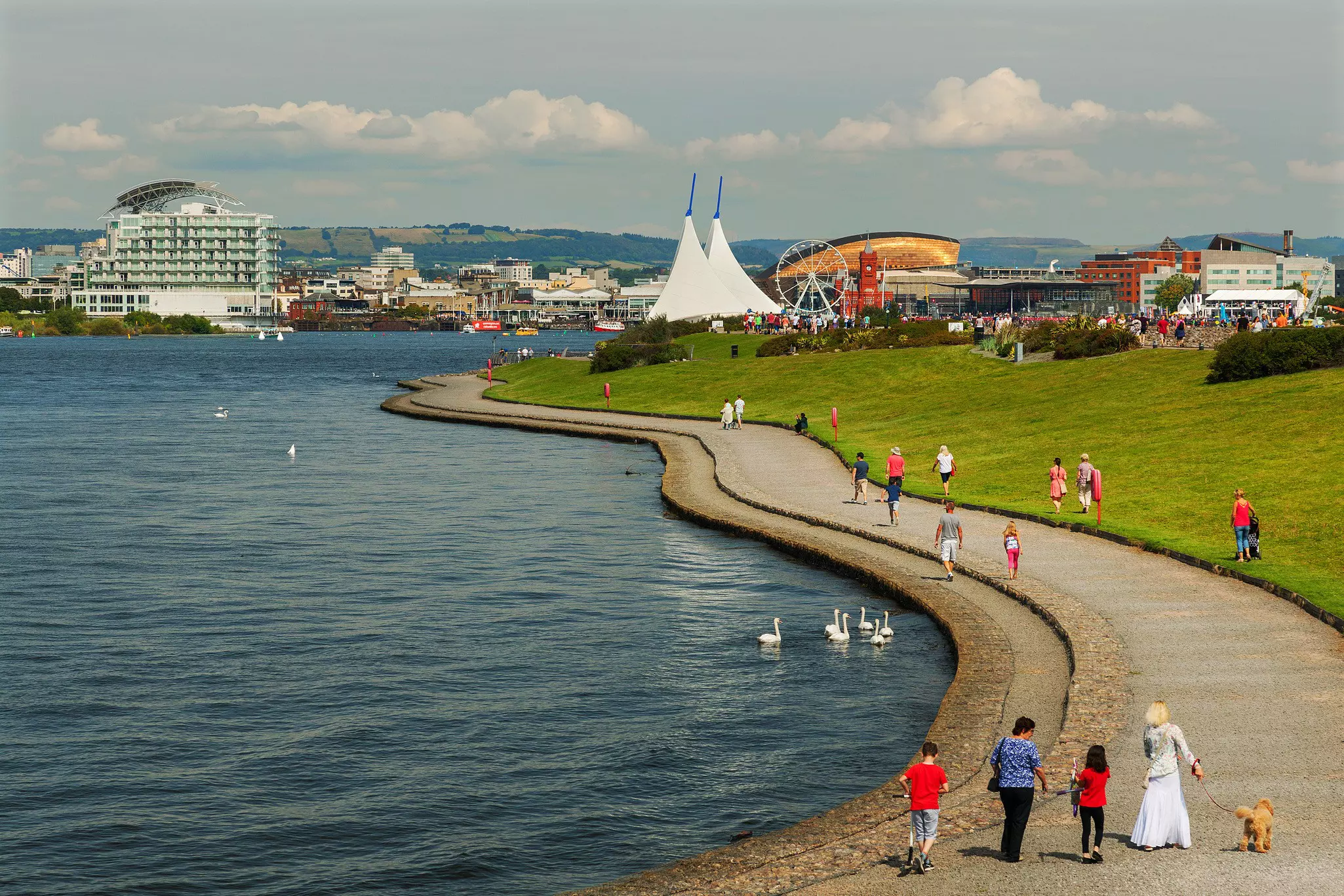 People walk along a waterside pathway leading towards a city dock with large buildings and a Ferris wheel