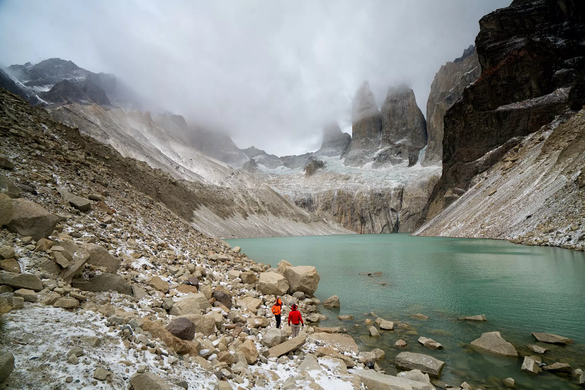 Two people in red jackets walk on a rocky hillside toward a clear blue lake surrounded by mountains; a low cloud hangs in the valley at the far shore.