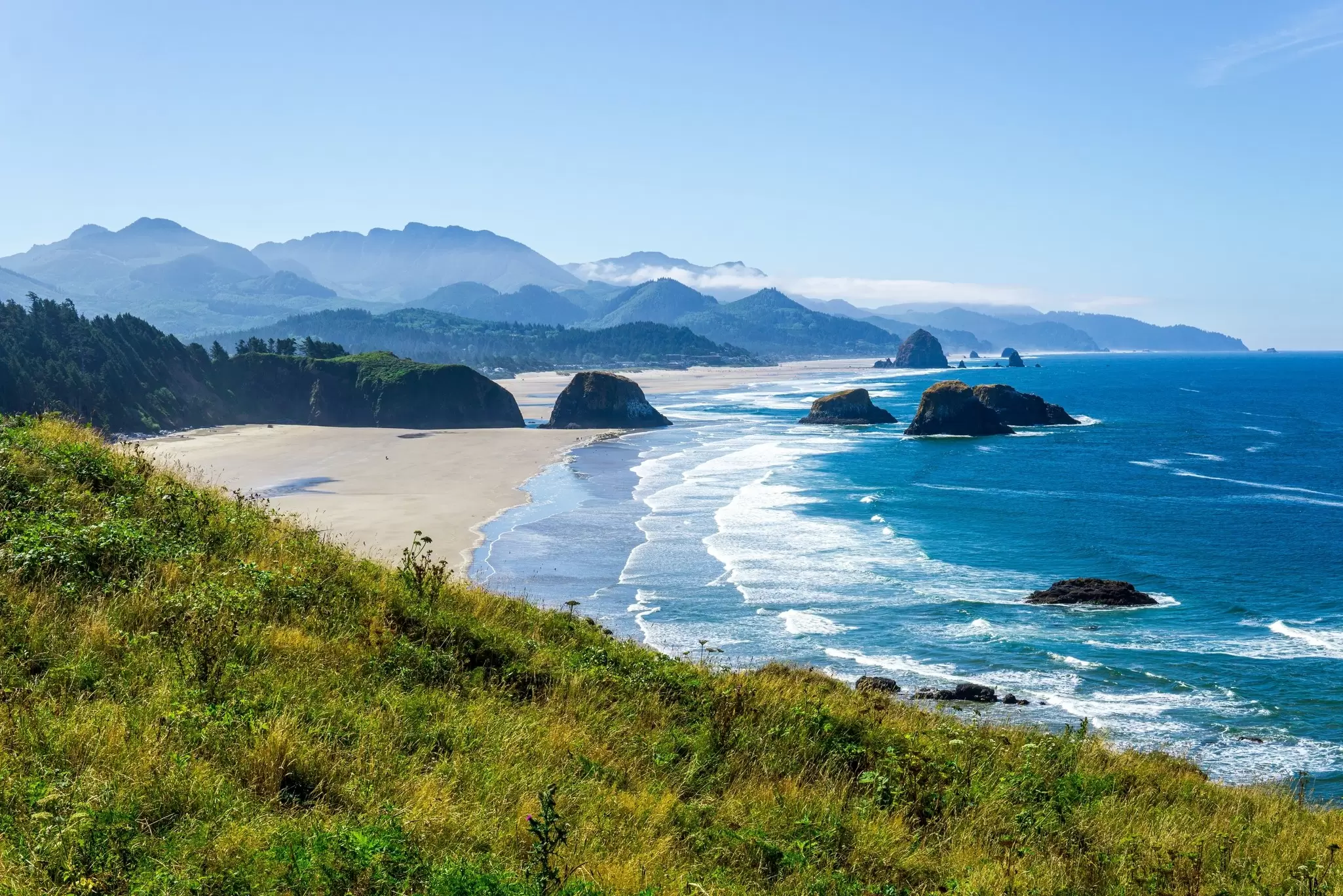 Blue ocean waves crash on a wide, sandy beach with sea stacks.