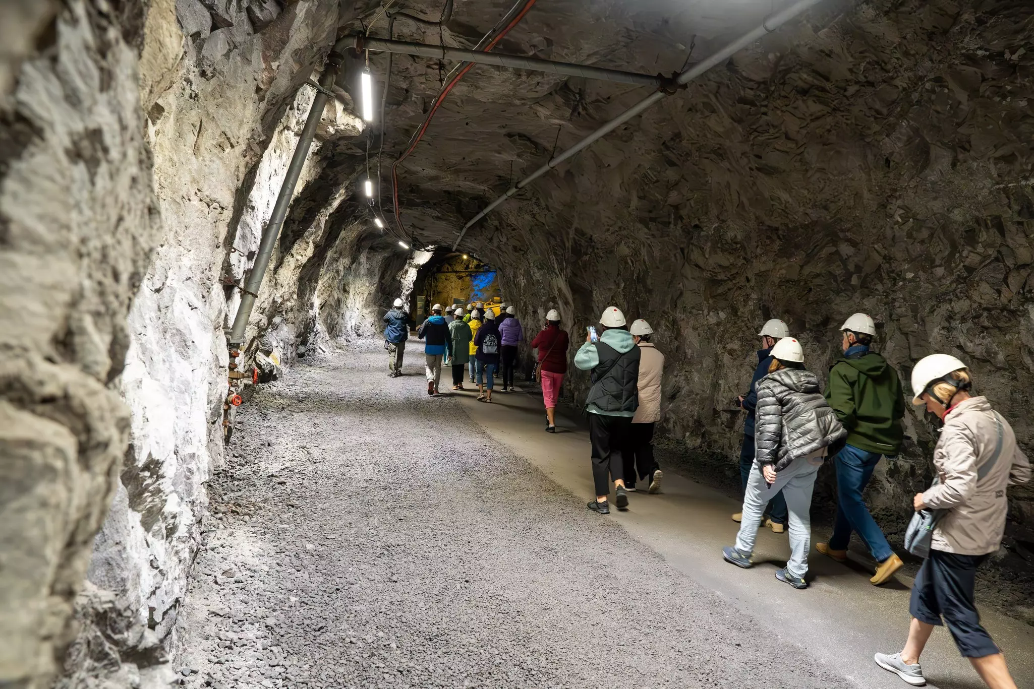 Visitors in hard hats walk through a wide tunnel in an underground mine.
