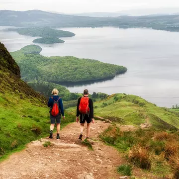 Hikers walk down a lush hill in front of a large lake.