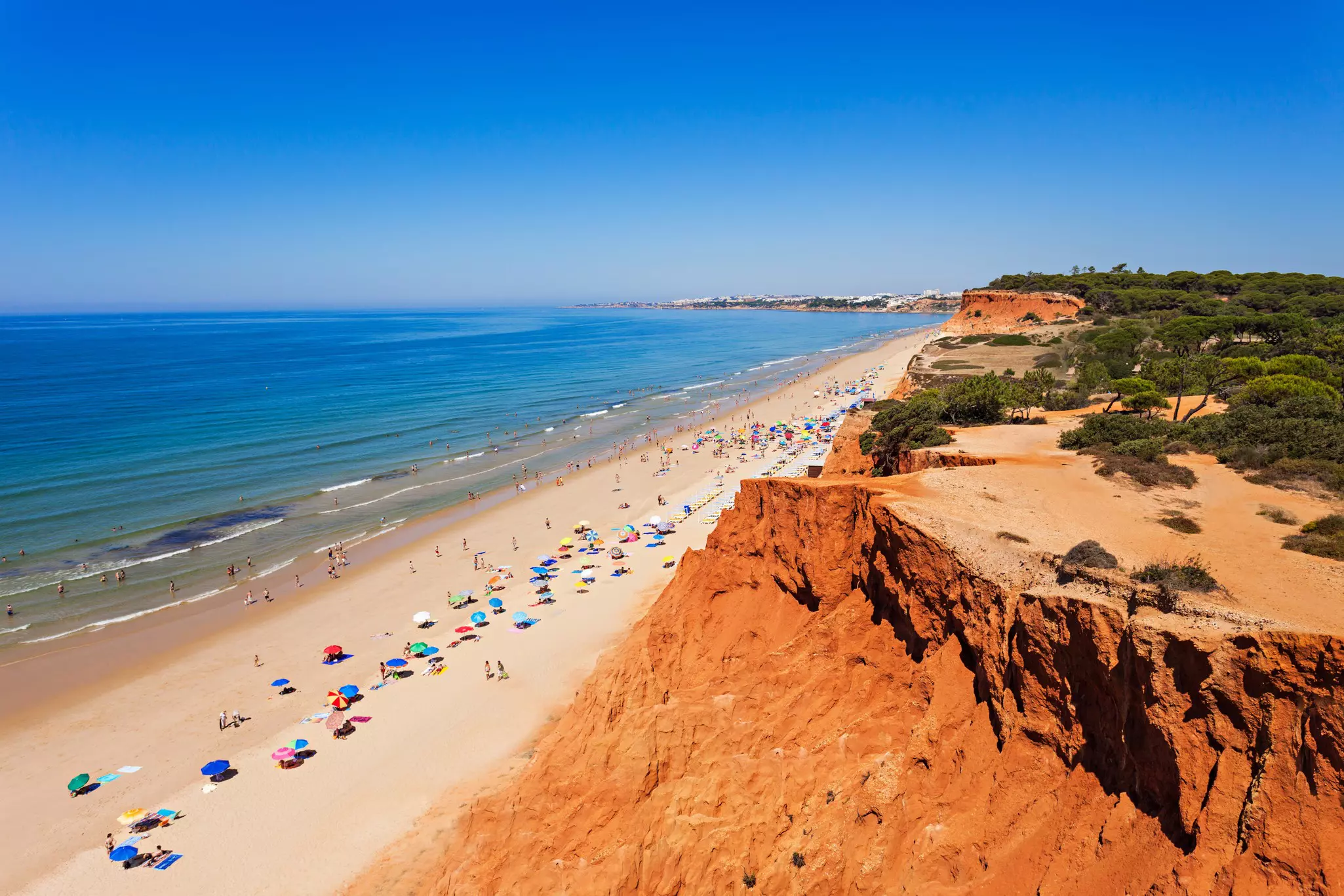 People under sun umbrellas on a long wide beach backed by red-sand cliffs.