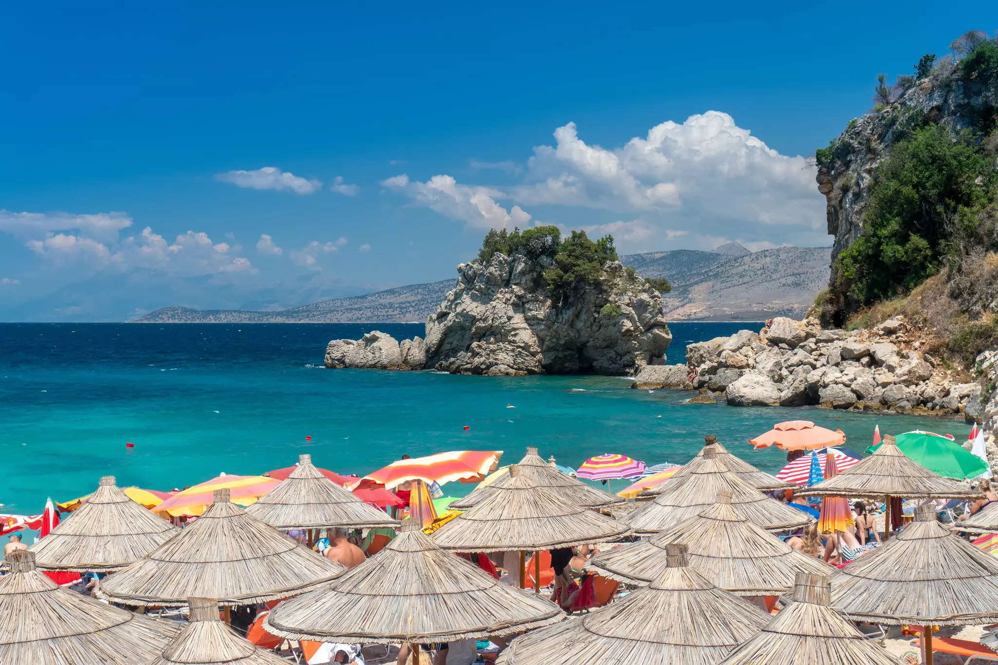 The tops of numerous straw sun umbrellas are pictured on beach. Turquoise-colored water and rocky islands are seen in the distance.