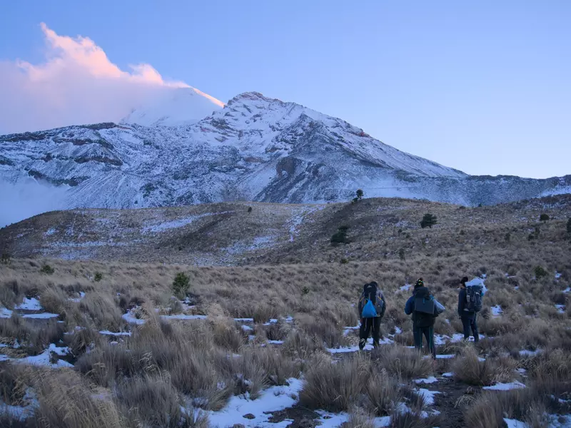 People hike across a snow-dusted field toward the snowy peak of a mountain at dawn.