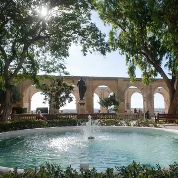 Round fountain pool in a shady garden, a bronze statue and a row of honey-coloured stone arches behind.
