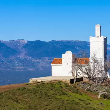 Whitewashed Spanish Mosque with a square minaret and terracotta roofs on a grassy hilltop, bare trees and mountains behind