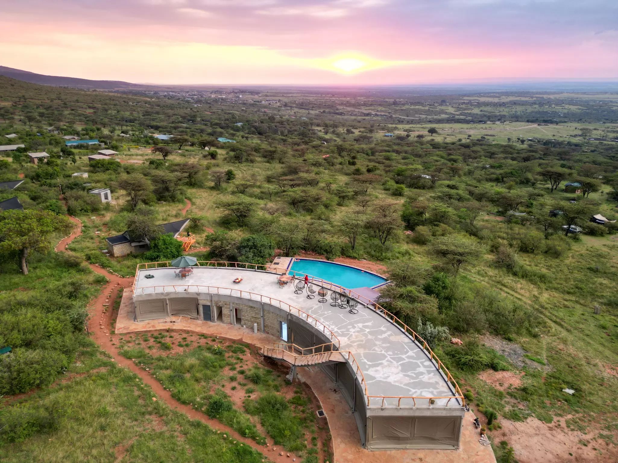 A curved building with a rooftop terrace and a rectangular swimming pool at the edge of a vast grassland at sunset.