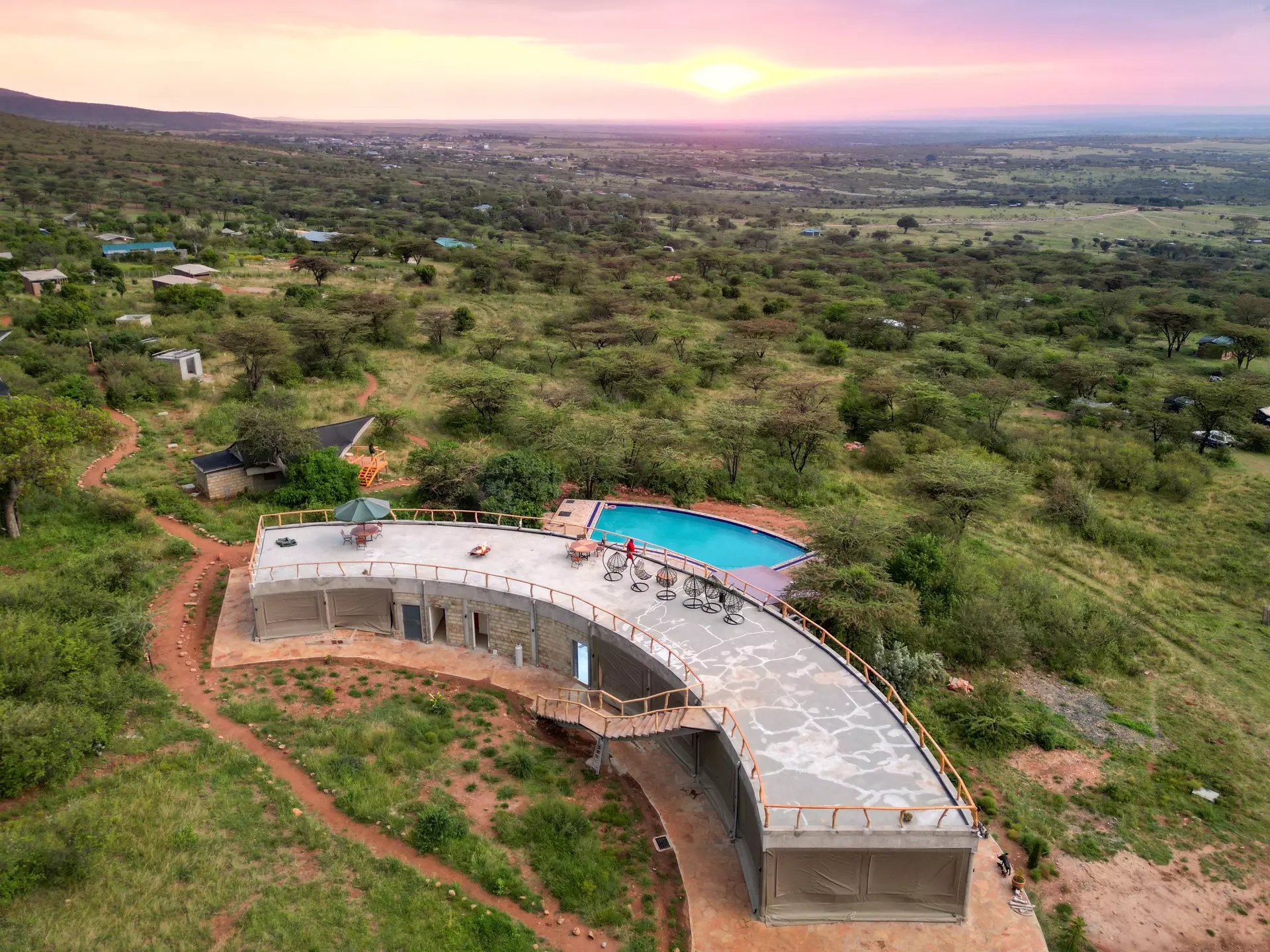 A curved building with a rooftop terrace and a rectangular swimming pool at the edge of a vast grassland at sunset.