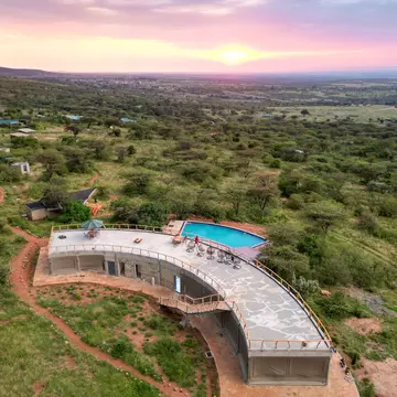 A curved building with a rooftop terrace and a rectangular swimming pool at the edge of a vast grassland at sunset.