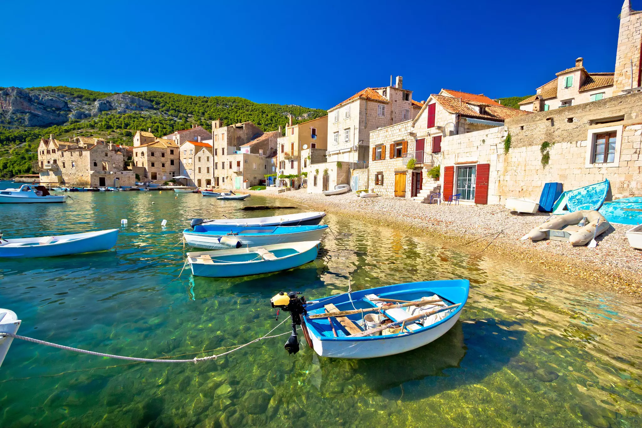 Small boats are moored in the harbor of a village, with stone houses lining the shore.
