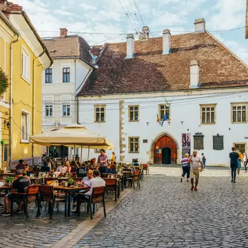 People dining at outdoor tables on a cobbled street in Cluj-Napoca, facing a white house with a Gothic stone doorway.