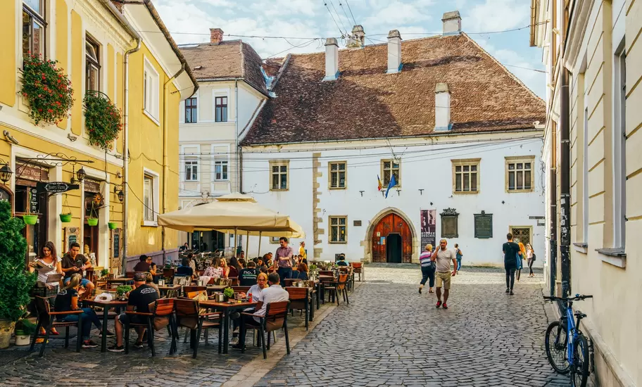 People dining at outdoor tables on a cobbled street in Cluj-Napoca, facing a white house with a Gothic stone doorway.