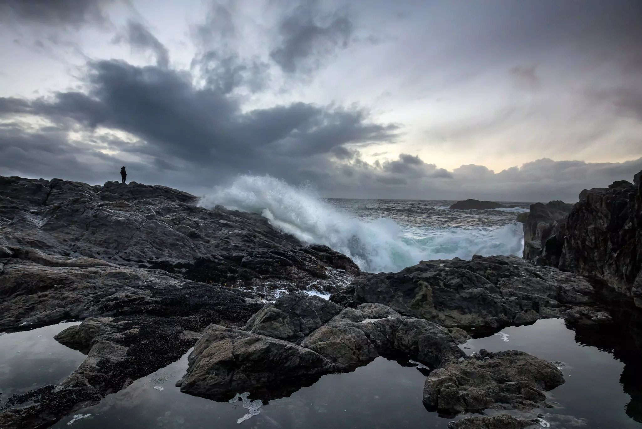A solo figure in silhouette stands on a rocky coastline watching dark storm clouds out at sea.