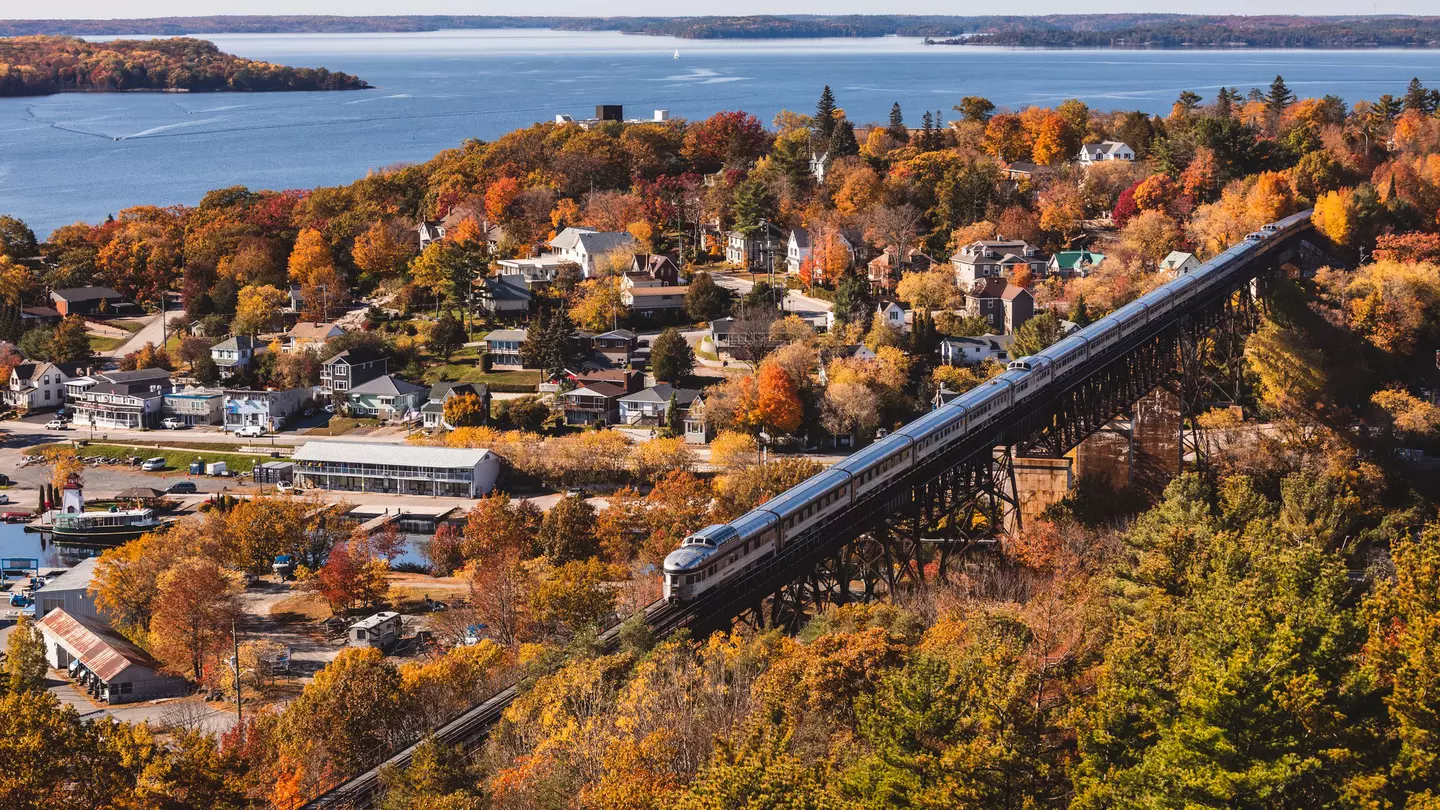 An aerial view of a train crossing a trestle by a charming waterfront town filled with trees in foliage