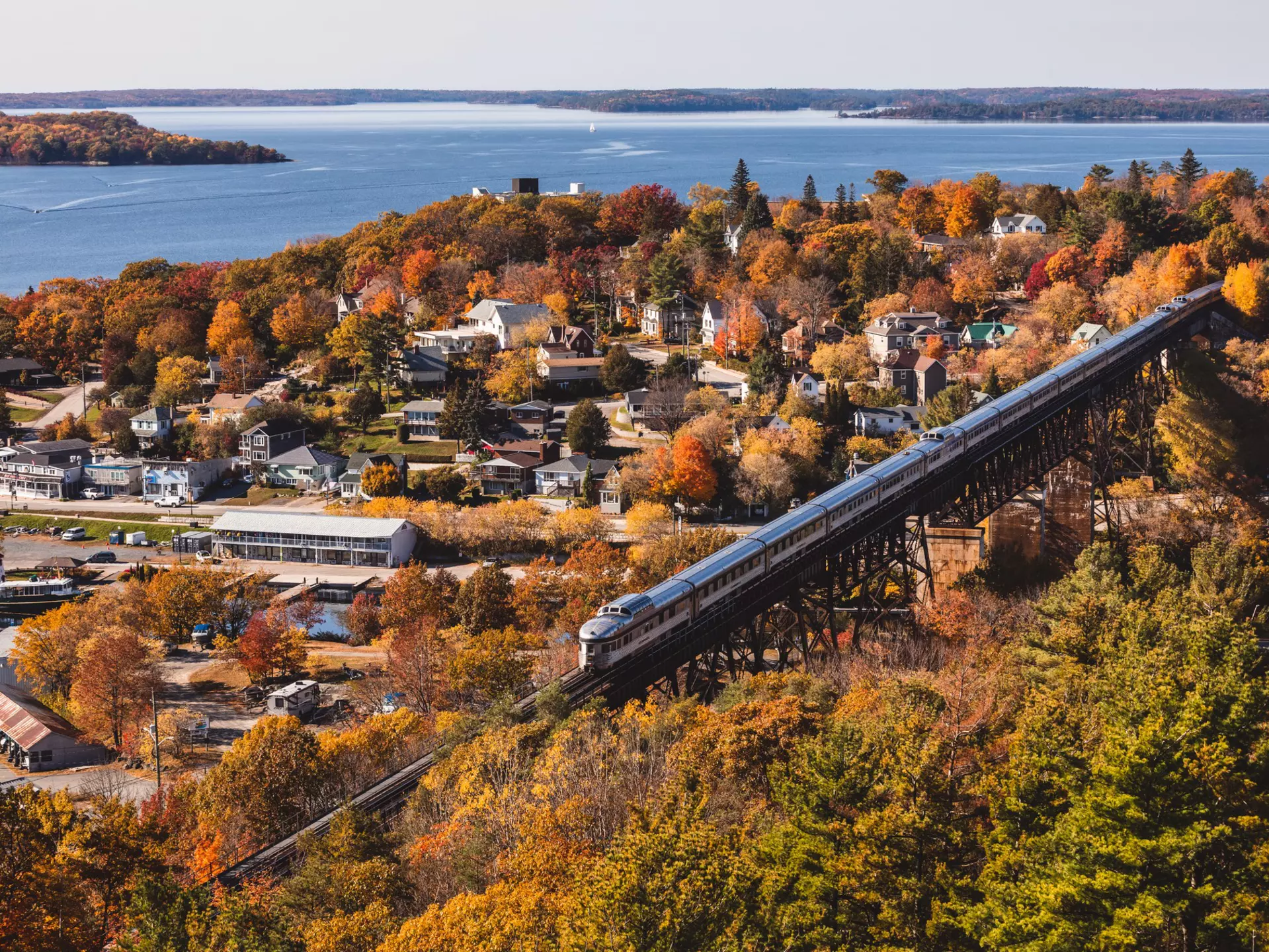 An aerial view of a train crossing a trestle by a charming waterfront town filled with trees in foliage