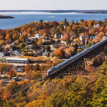 An aerial view of a train crossing a trestle by a charming waterfront town filled with trees in foliage