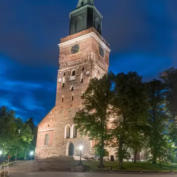 The brick tower of Turku Cathedral, floodlit at night beneath a deep blue sky, with trees and an empty cobbled square.