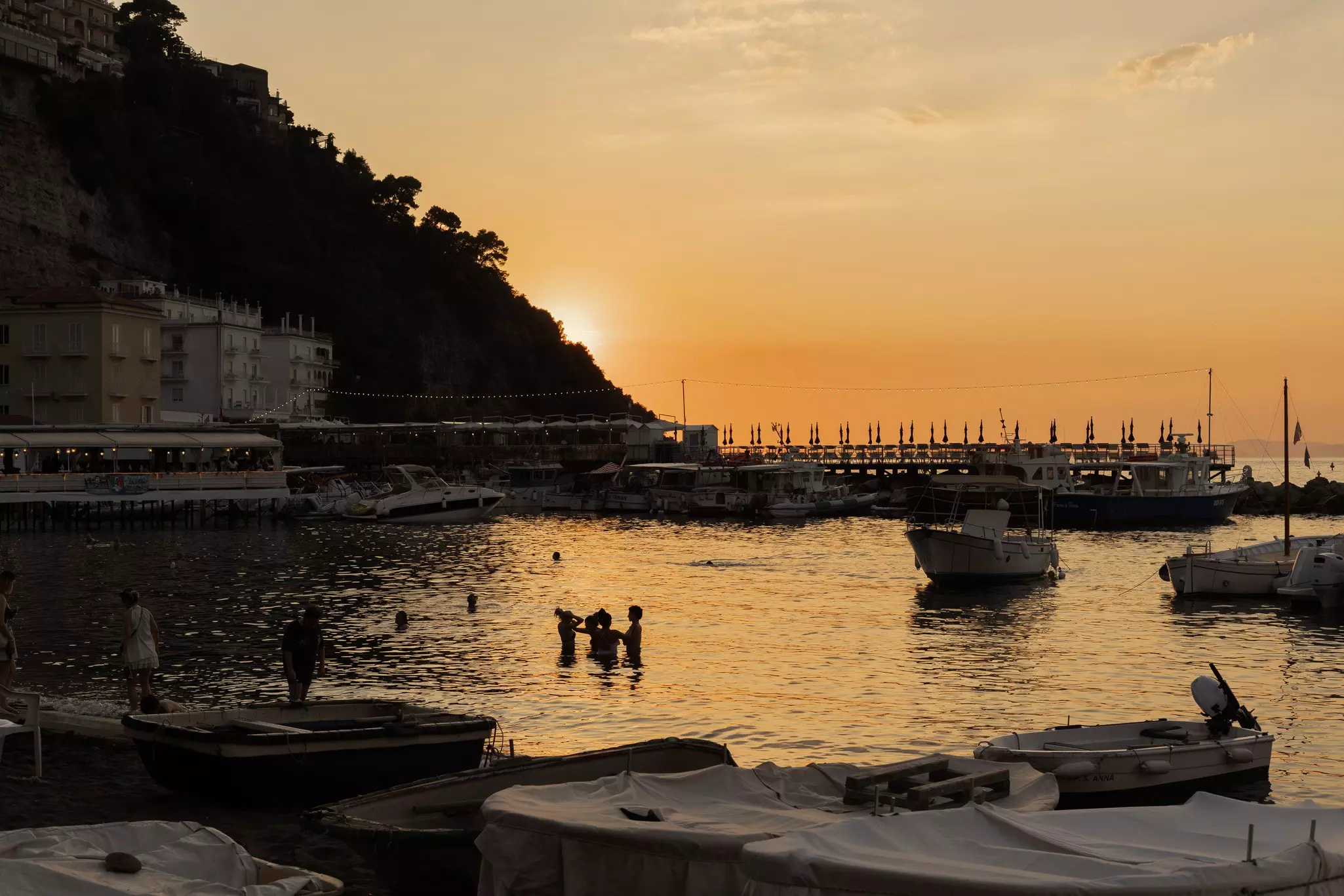 Silhouettes of people in a body of water near boats and a dock at sunset