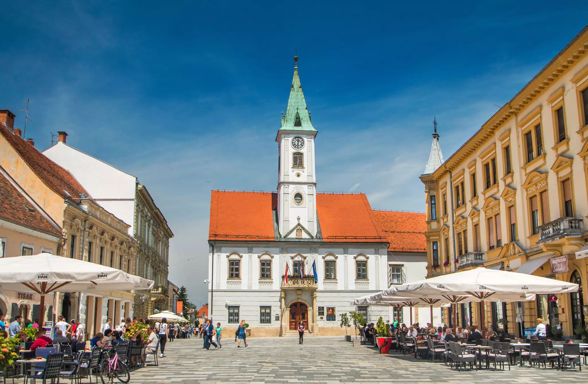 A square in Croatia has cafes with outdoor tables under umbrellas on either side and a white building with a steeple and red roof at one end.