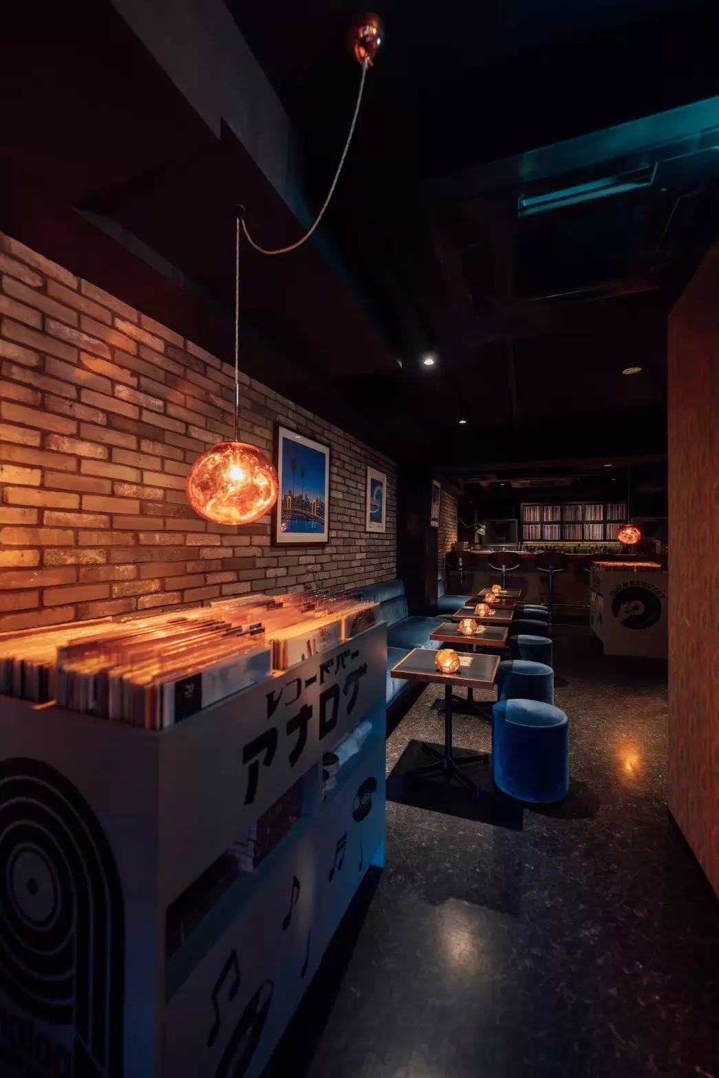 A box of records stands in front of a short row of seating and tables. A bar at the back of the room is backed by a vast collection of records.