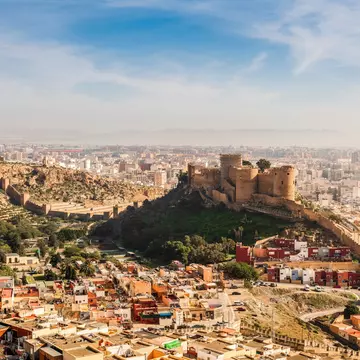 A vast ancient fortress citadel on a hill above a coastal town.