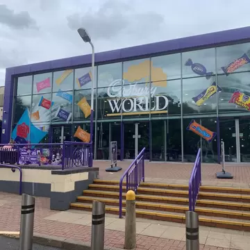 Purple-framed glass building entrance with 'Cadbury World' lettering and large chocolate bar graphics, reached by steps.
