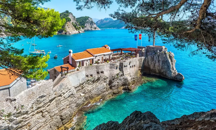 Venetian fortress on a rocky point in Petrovac, Montenegro, with flags flying over turquoise water and moored boats