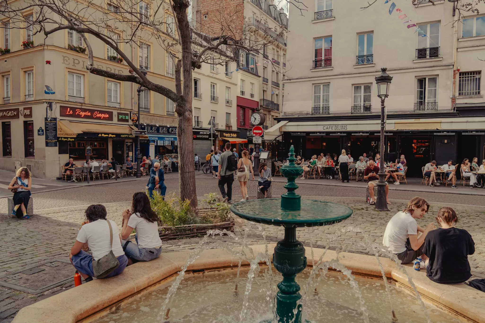 People sit on the edge of a fountain in a city square, people-watching. Other people eat on restaurant terraces in the background