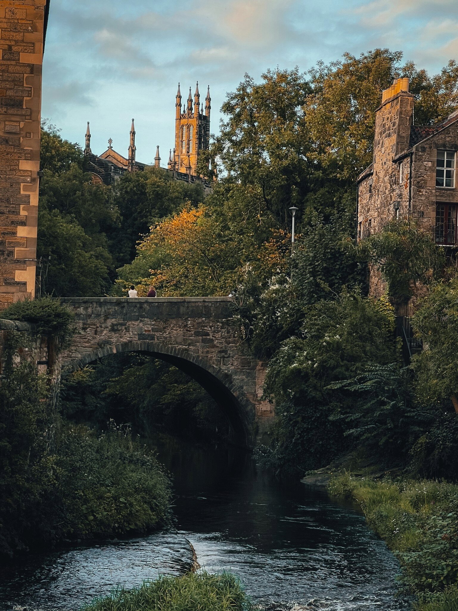 A peaceful stone bridge crossing a river surrounded by lush greenery, with a historic church and spires in the background at sunset in Dean Village, Edinburgh.