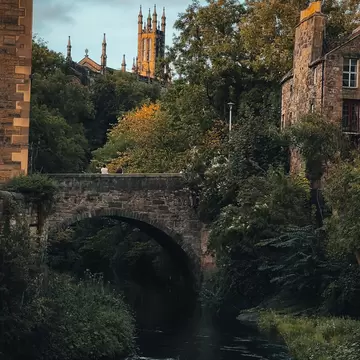 A peaceful stone bridge crossing a river surrounded by lush greenery, with a historic church and spires in the background at sunset in Dean Village, Edinburgh.