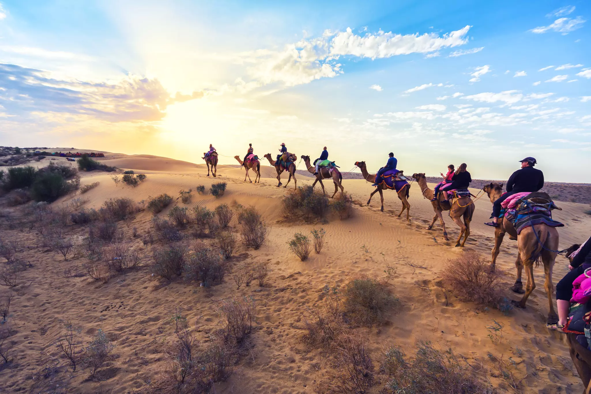 A caravan of camels carrying riders processes through a desert at sunset.