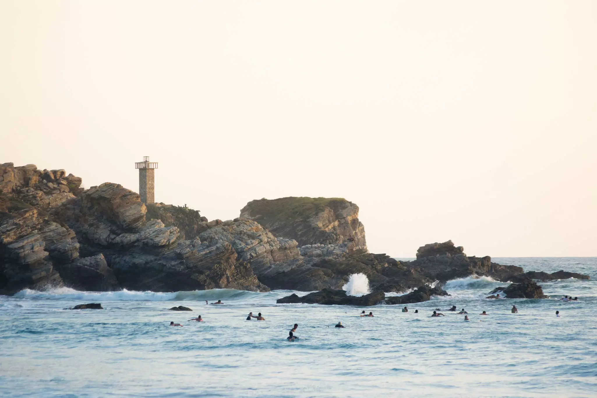 Evening light paints the lighthouse of Punta Zicatela, while many surfers pack the lineup in Puerto Escondido, Mexico