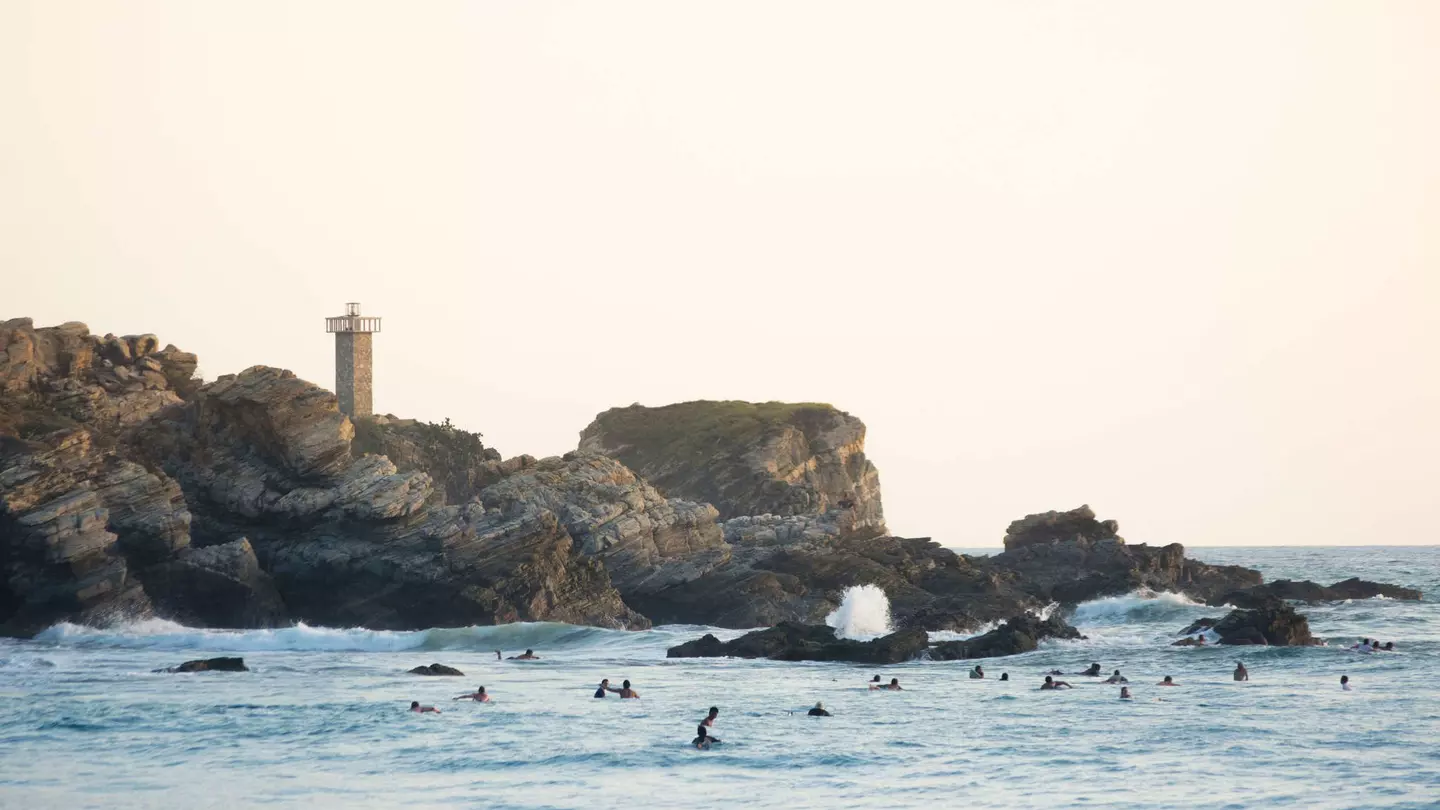 Evening light paints the lighthouse of Punta Zicatela, while many surfers pack the lineup in Puerto Escondido, Mexico
