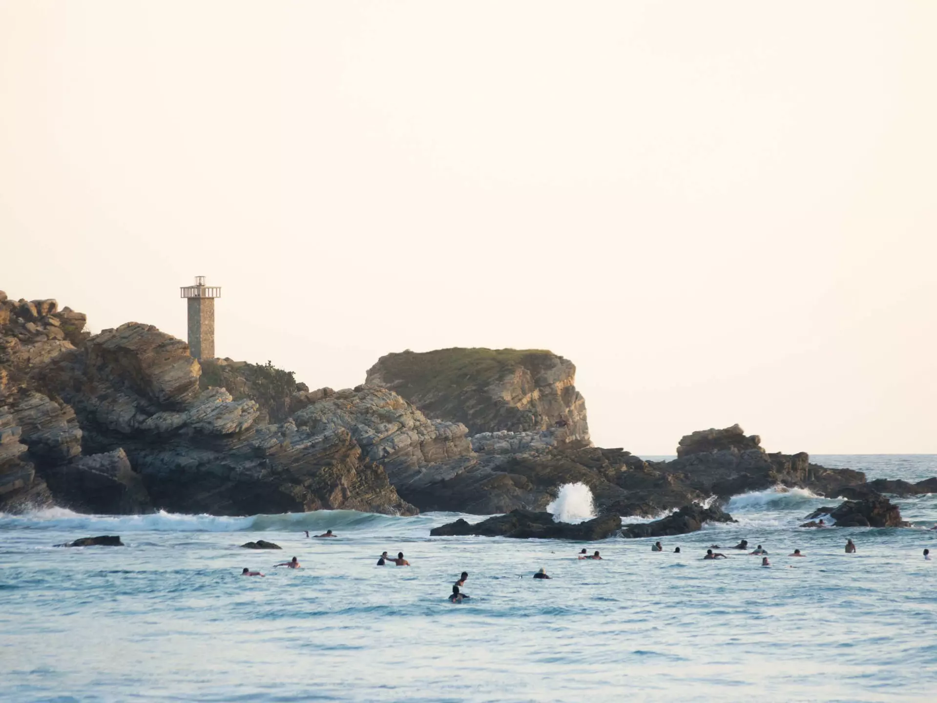 Evening light paints the lighthouse of Punta Zicatela, while many surfers pack the lineup in Puerto Escondido, Mexico