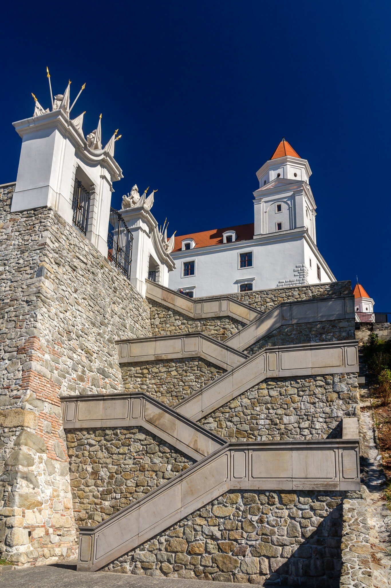 A wide stone stairway zigzagging up a rubble rampart towards a white castle with red-roofed corner towers, under a deep blue sky.