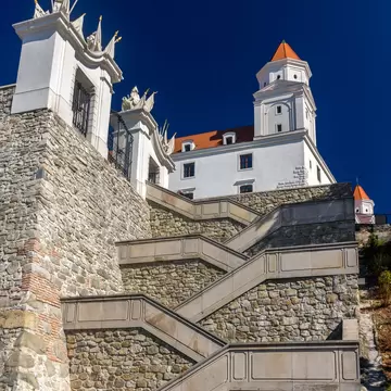 A wide stone stairway zigzagging up a rubble rampart towards a white castle with red-roofed corner towers, under a deep blue sky.