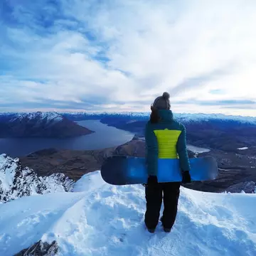 A woman holding a snowboard at the top of a snowy mountain at the Remarkables Ski Area