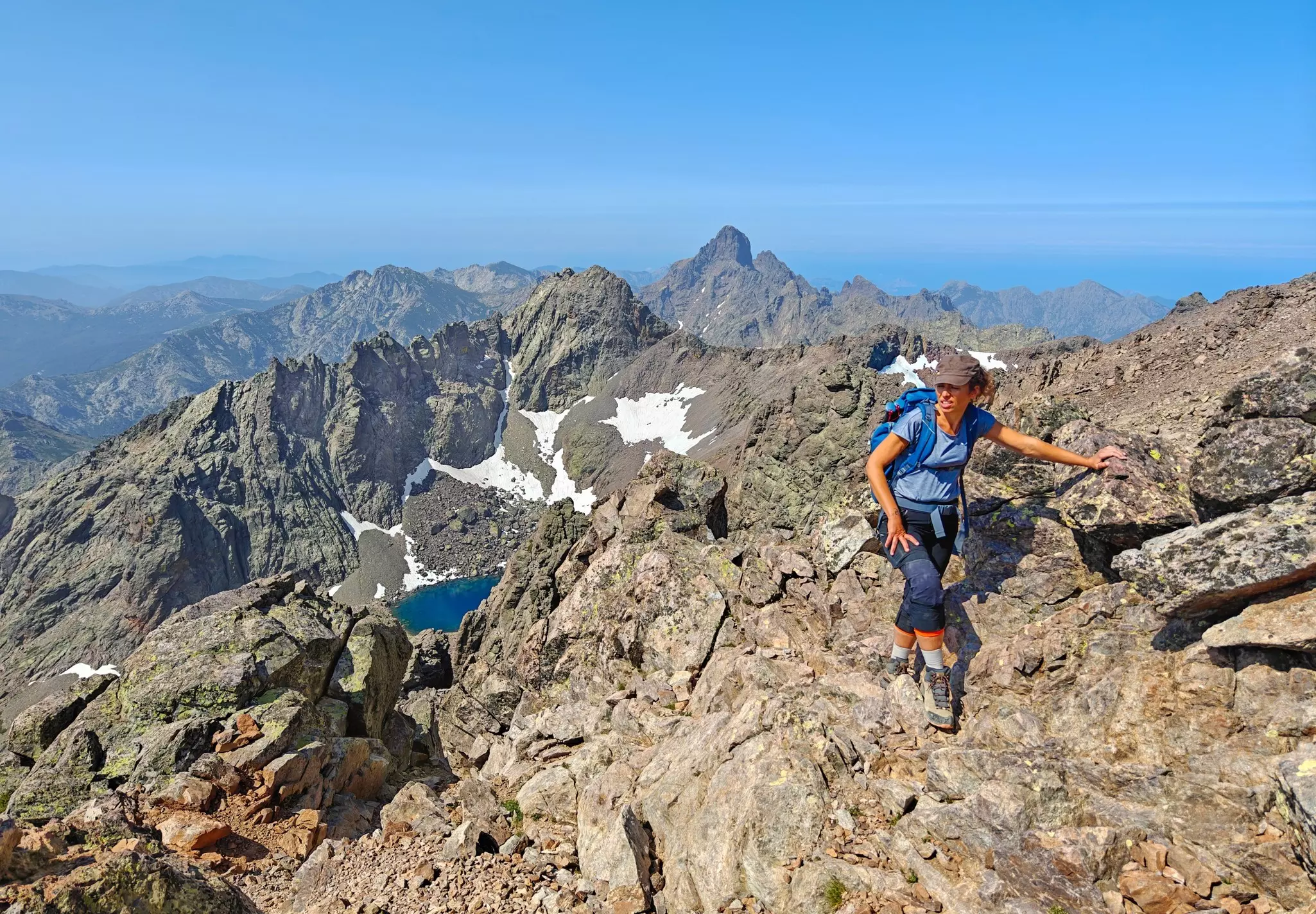 A view of a female hiker at the peak of a rocky mountain. Other rocky hills are visible in the distance, as is the sea.